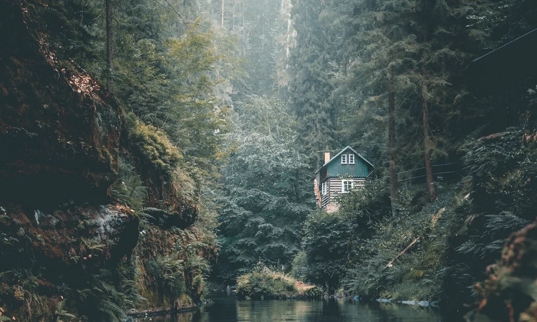 A log cabin in the woods with a still water source nearby. Moss gathers on the rocks rising out of the river.